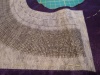 A piece of purple sateen fabric topped by a stabiliser printed with a curved grid. Blackwork-style embroidery in a silver thread has been started on the grid.