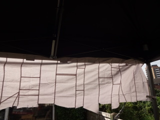 A short grey/silver curtain hanging from a gazebo, with the sun behind it. It's made from small rectangles of fabric, and the seam allowances show up as darker lines.
